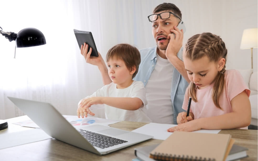 Far sidder ved skrivebordet med sine to børn og forsøger at arbejde, mens han passer dem.