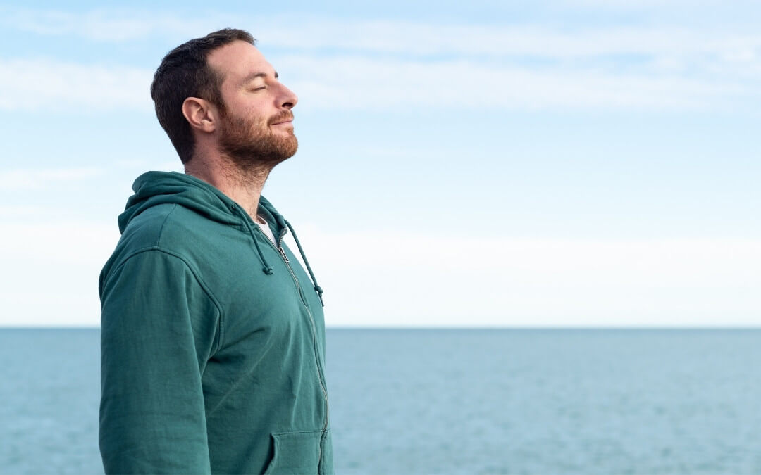 Mand står på stranden med lukkede øjne og et stille smil, opslugt af sine tanker.